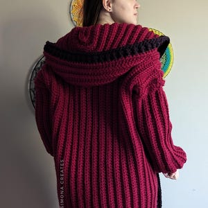 Pu&ograve; includere: Un cardigan in crochet color borgogna con un bordo nero intorno al cappuccio e sui lati. Il cardigan ha una trama a coste e una vestibilit&agrave; lunga e ampia.