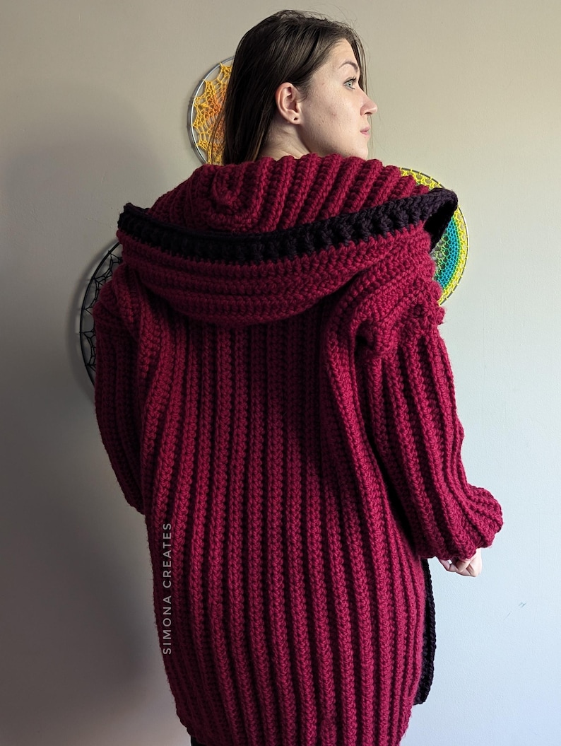 Pu&ograve; includere: Un cardigan in crochet color borgogna con un bordo nero intorno al cappuccio e sui lati. Il cardigan ha una trama a coste e una vestibilit&agrave; lunga e ampia.