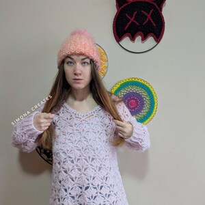 May include: A pink crocheted sweater with a lacy pattern. The sweater has long sleeves and a loose fit. The model is wearing a pink and orange beanie with a fluffy pom-pom.