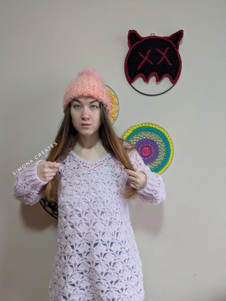 May include: A pink crocheted sweater with a lacy pattern. The sweater has long sleeves and a loose fit. The model is wearing a pink and orange beanie with a fluffy pom-pom.