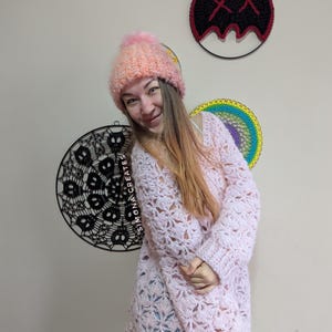 May include: A woman wearing a pink crocheted sweater and a pink and white crocheted hat with a pom pom. She is standing in front of a wall with two macrame wall hangings.