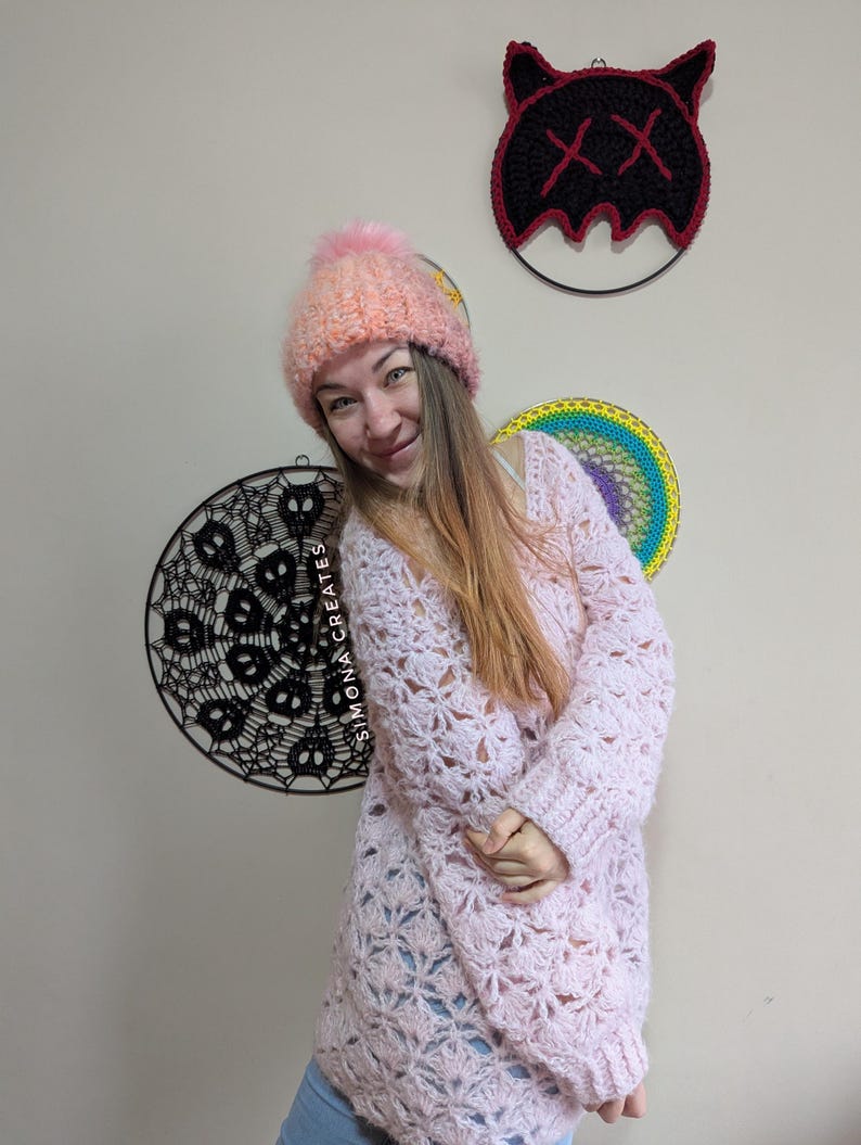 May include: A woman wearing a pink crocheted sweater and a pink and white crocheted hat with a pom pom. She is standing in front of a wall with two macrame wall hangings.
