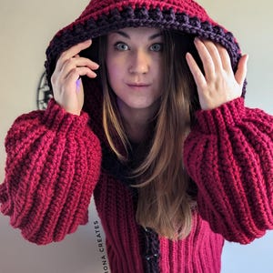 Pu&ograve; includere: Un cardigan con cappuccio in crochet rosso con un bordo viola scuro. Il cardigan ha una chiusura a bottone e una trama a coste.