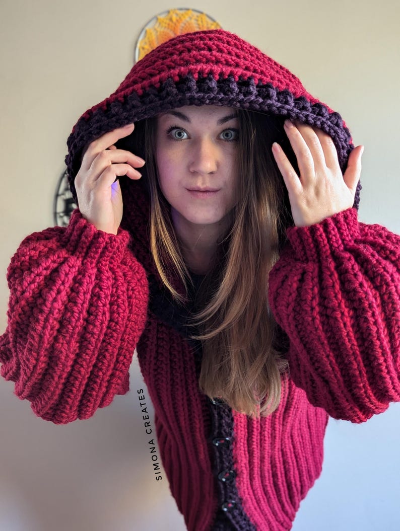 Pu&ograve; includere: Un cardigan con cappuccio in crochet rosso con un bordo viola scuro. Il cardigan ha una chiusura a bottone e una trama a coste.
