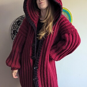 Pu&ograve; includere: Un lungo cardigan in crochet color borgogna con bordino nero e cappuccio. Il cardigan ha una chiusura con bottoni ed &egrave; realizzato con un filato spesso.