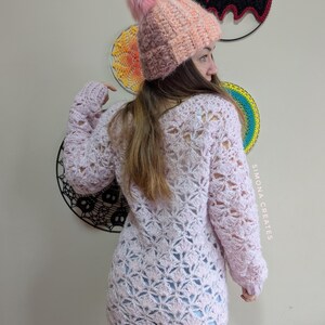 May include: A pink crocheted sweater with a lacy pattern. The sweater has long sleeves and a loose fit. The model is wearing a pink and orange crocheted hat with a fluffy pom-pom.