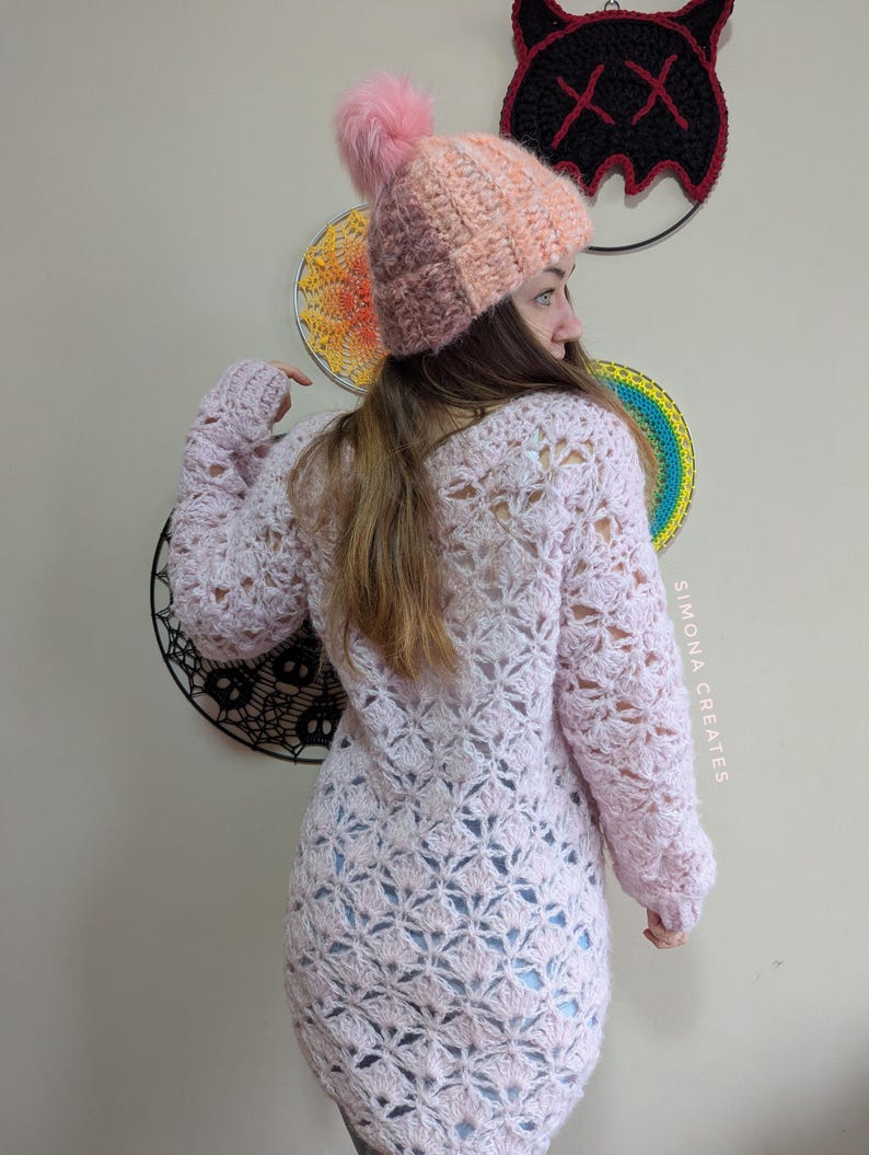 May include: A pink crocheted sweater with a lacy pattern. The sweater has long sleeves and a loose fit. The model is wearing a pink and orange crocheted hat with a fluffy pom-pom.