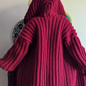 Pu&ograve; includere: Un cardigan di uncinetto rosso scuro con un grande cappuccio. Il cardigan &egrave; realizzato con un filato spesso e ha una trama a coste.