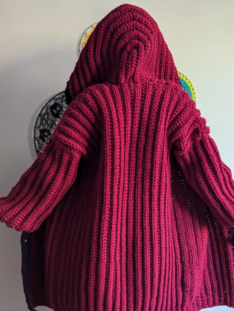 Pu&ograve; includere: Un cardigan di uncinetto rosso scuro con un grande cappuccio. Il cardigan &egrave; realizzato con un filato spesso e ha una trama a coste.