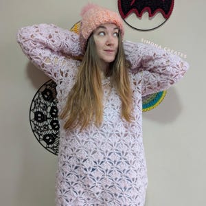 May include: A woman wearing a pink crocheted sweater with a white crocheted hat. The sweater has a lacy pattern and the hat has a fluffy pom-pom. The woman is standing in front of a wall with a black and red crocheted wall hanging.
