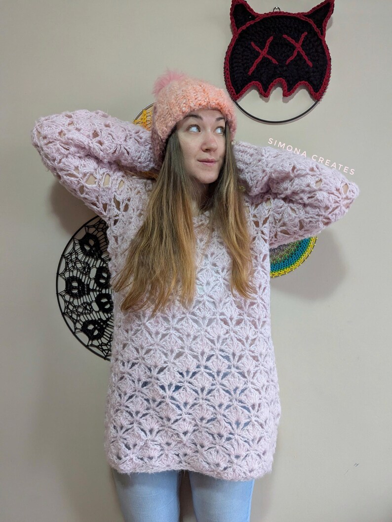 May include: A woman wearing a pink crocheted sweater with a white crocheted hat. The sweater has a lacy pattern and the hat has a fluffy pom-pom. The woman is standing in front of a wall with a black and red crocheted wall hanging.