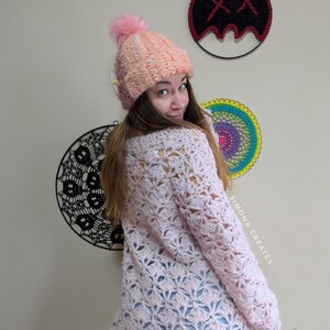 May include: A pink crocheted sweater with a lacy pattern. The sweater is worn by a person with a pink and white crocheted hat with a fluffy pom pom. The text "SIMONA CREATES" is visible on the right side of the image.