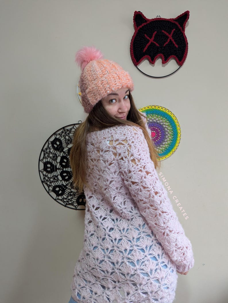 May include: A pink crocheted sweater with a lacy pattern. The sweater is worn by a person with a pink and white crocheted hat with a fluffy pom pom. The text "SIMONA CREATES" is visible on the right side of the image.