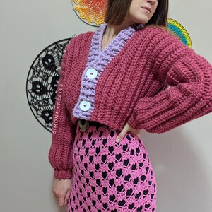 Pu&ograve; includere: Un cardigan in crochet rosa e viola con chiusura a bottone. Il cardigan &egrave; corto e ha una trama a coste. La modella indossa una gonna in crochet rosa con un motivo a forma di cuore.