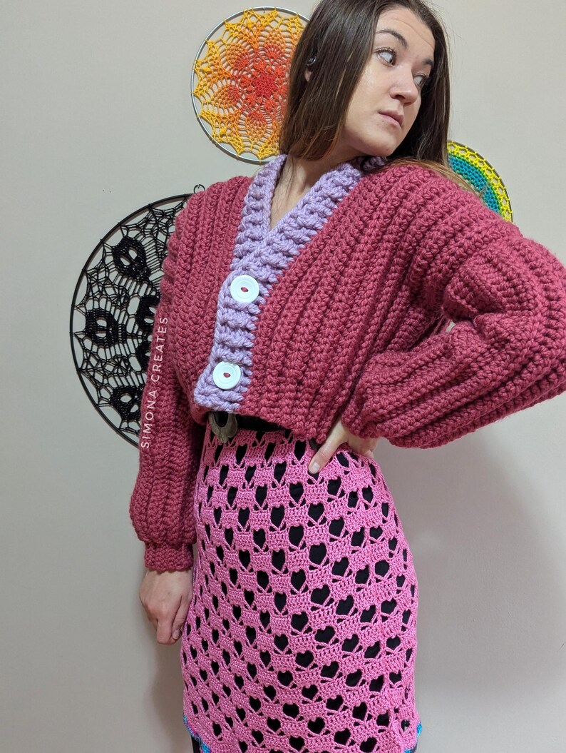 Pu&ograve; includere: Un cardigan in crochet rosa e viola con chiusura a bottone. Il cardigan &egrave; corto e ha una trama a coste. La modella indossa una gonna in crochet rosa con un motivo a forma di cuore.