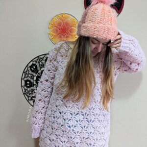 May include: A pink crocheted sweater with a lacy pattern and long sleeves. The sweater is worn over blue jeans and paired with a pink crocheted beanie with a fluffy pom-pom.