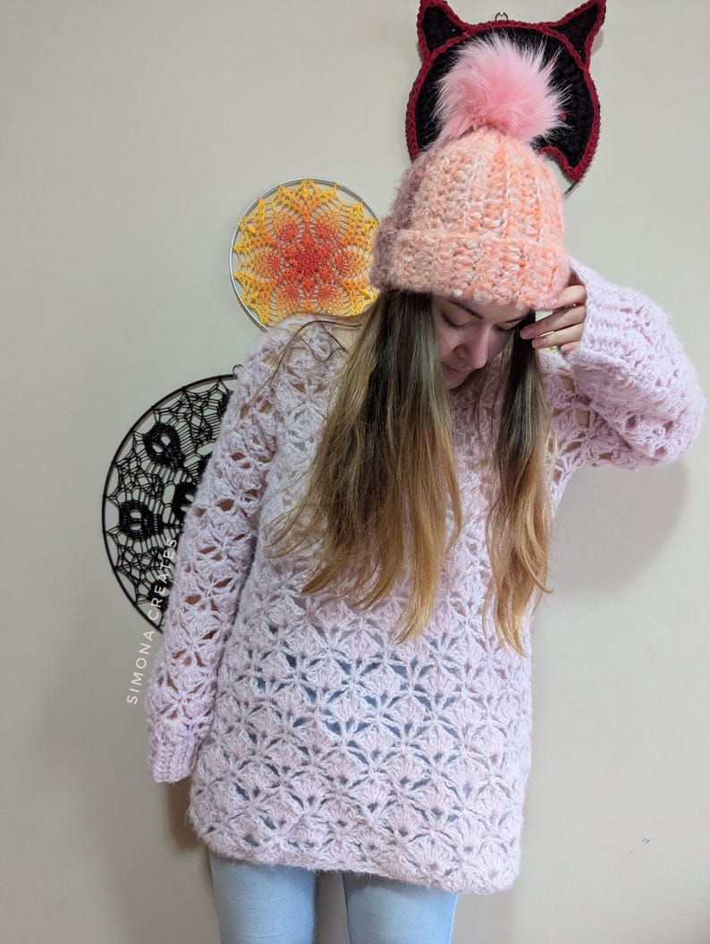 May include: A pink crocheted sweater with a lacy pattern and long sleeves. The sweater is worn over blue jeans and paired with a pink crocheted beanie with a fluffy pom-pom.