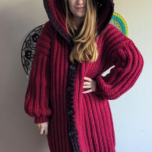 Pu&ograve; includere: Un lungo cardigan rosso all'uncinetto con cappuccio e bordi neri. Il cardigan ha una chiusura con bottoni ed &egrave; realizzato con un filato spesso.
