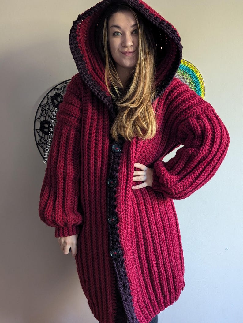 Pu&ograve; includere: Un lungo cardigan rosso all'uncinetto con cappuccio e bordi neri. Il cardigan ha una chiusura con bottoni ed &egrave; realizzato con un filato spesso.