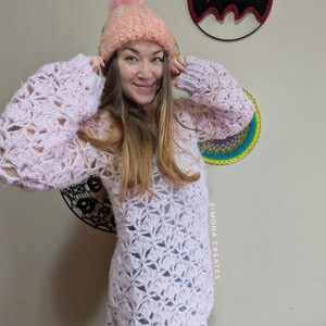 May include: A pink crocheted sweater with a lacy pattern and a white crocheted hat with a fluffy pom pom. The sweater has long sleeves and a loose fit. The hat is pulled down over the person's ears. The text "SIMONA CREATES" is visible on the sweater.