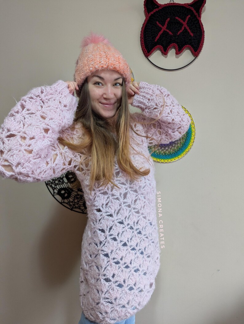May include: A pink crocheted sweater with a lacy pattern and a white crocheted hat with a fluffy pom pom. The sweater has long sleeves and a loose fit. The hat is pulled down over the person's ears. The text "SIMONA CREATES" is visible on the sweater.