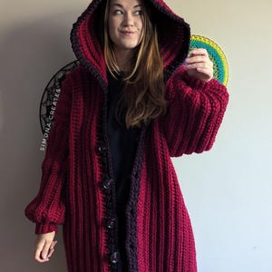 Pu&ograve; includere: Un lungo cardigan in crochet color borgogna con bordino nero e cappuccio. Il cardigan ha una chiusura con bottoni ed &egrave; realizzato con un filato spesso.