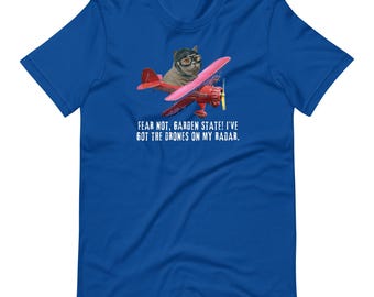 May include: Royal blue t-shirt featuring a cat wearing aviator goggles and a cap, piloting a red biplane with pink wings. The text on the shirt reads: "FEAR NOT, GARDEN STATE! I'VE GOT THE DRONES ON MY RADAR."