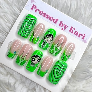May include: Green and pink press-on nails with a green and black cartoon character design. The nails feature a heart design, flames, and glitter. The text "Pressed by Kari" is on the white card.