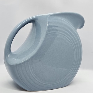 May include: A light blue ceramic pitcher with a rounded shape and a wide, flared spout. The pitcher has a smooth, glossy finish and a subtle ribbing pattern.