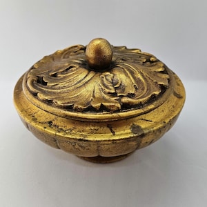 May include: A gold-coloured decorative trinket box with an ornate, carved lid and a round knob. The box has a distressed, aged look.