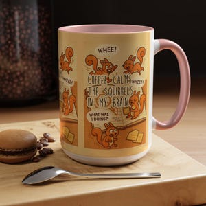 May include: A white ceramic coffee mug with a pink rim and handle. The mug features a cartoon illustration of squirrels and the text "WHEEE! COFFEE CALMS THE SQUIRRELS IN MY BRAIN WHEE! WHAT WAS I DOING?"