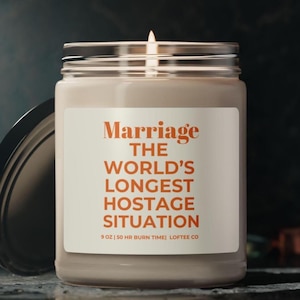 May include: A white candle jar with a label that reads "Marriage The World's Longest Hostage Situation" in orange text. The candle is 9 oz and has a 50 hour burn time. The label also includes the text "Loftee Co."