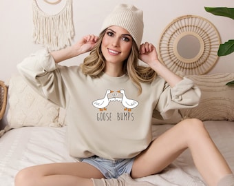 May include: A light beige sweatshirt featuring a graphic of two geese facing each other with the text "Goose Bumps" underneath. The model is wearing the sweatshirt and a white beanie.