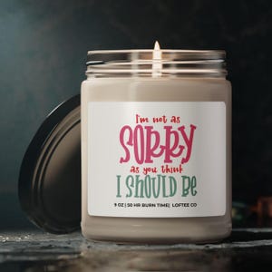 May include: A beige candle jar with a white label that reads "I'm not as SORRY as you think I SHOULD BE". The label also includes "9 OZ | 50 HR BURN TIME | LOFTEE CO".
