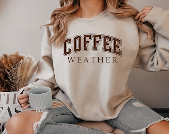 May include: A beige sweatshirt with the text "COFFEE WEATHER" in brown letters. The person is holding a light gray mug.