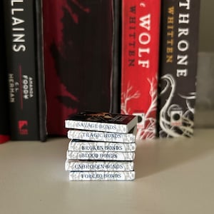 Puede incluir: Una pila de seis libros de bolsillo con cubiertas blancas y títulos azules. Los títulos son "Savage Bonds", "Tragic Bonds", "Broken Bonds", "Blood Bonds", "Unbroken Bonds" y "Forced Bonds".