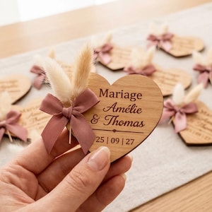 Puede incluir: Recuerdo de boda de madera en forma de corazón con un lazo malva y acentos florales secos. El recuerdo está grabado con los nombres Amélie & Thomas y la fecha 25/09/27. Otros recuerdos son visibles en el fondo.