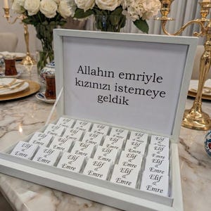 May include: A white box filled with small chocolate bars, each wrapped in white paper with the names "Elif & Emre" printed on them. The box lid has text in Turkish. White roses and gold candle holders are in the background.