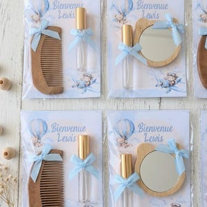 Puede incluir: Recuerdos de baby shower en envases de plástico transparente. Cada recuerdo incluye un peine de madera, un pequeño frasco de perfume con tapa dorada o un pequeño espejo de madera. Todos están atados con una cinta azul claro. El texto "Bienvenue Lewis".