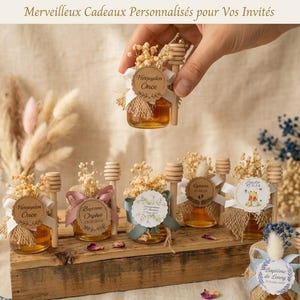Könnte beinhalten: Kleine Honiggläser aus Glas, verziert mit Juteband, weißer Schleife, Trockenblumen und einem hölzernen Honiglöffel. Jedes Glas ist mit einem personalisierten runden Etikett versehen, das Namen und Daten trägt und auf einem Holzbrett steht.