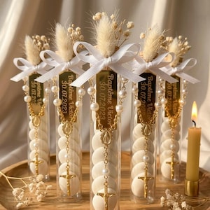 May include: Clear tubes filled with white wafers, adorned with gold rosaries, and topped with dried flowers and white ribbons. Each tube has a gold tag with the text "Baptême Victor 30.07.2023". A lit candle sits nearby on a wooden tray.