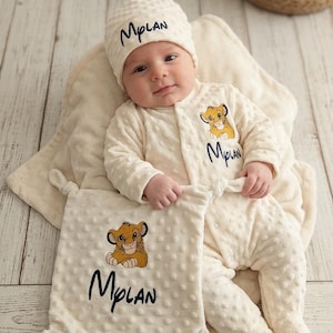 Puede incluir: Un bebé con un conjunto, gorro y manta a juego de color crema. El conjunto y la manta presentan un león de dibujos animados y el nombre "Mplan" en escritura negra. El bebé está acostado sobre una manta crema.