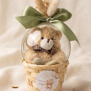 May include: A teddy bear in a clear glass sphere, decorated with a green ribbon and dried flowers, sits on a woven basket. The basket features a sticker with a teddy bear illustration and the text "Bienvenue Noah".
