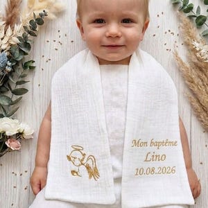 Può includere: Un bavaglino bianco per neonati con ricamo dorato. Presenta un angelo e la scritta "Mon baptême Lino 10.08.2026". Il bambino indossa una tutina bianca. Fiori secchi sullo sfondo.