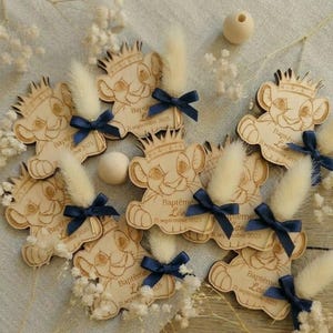 Può includere: Bomboniere a forma di cucciolo di leone in legno con corone, fiocchi blu navy e accenti bianchi soffici. Ogni bomboniera ha il testo "Baptême Léo 21 settembre 2025" inciso. Le bomboniere sono disposte con fiori bianchi e perline di legno.