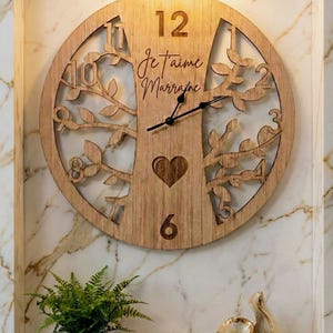 Peut inclure: Horloge murale en bois avec un motif d'arbre, avec les chiffres 12, 1, 2, 3, 6, 9, 10 et 11. L'horloge a une forme de cœur et les mots "Je t'aime Marraine". L'horloge est accrochée à un mur en marbre.