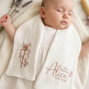 Può includere: Un bambino che dorme indossa una sciarpa battesimale in mussola bianca con dettagli ricamati. La sciarpa presenta un disegno a croce e floreale, insieme al nome "Alice" e alla data 12.06.2028.