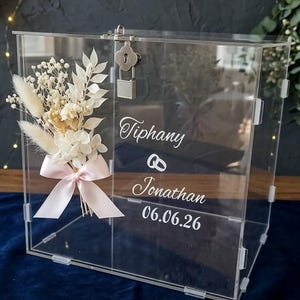 Könnte beinhalten: Transparente Hochzeitskartenbox aus Acryl mit Schloss und Schlüssel. Verziert mit einem Strauß Trockenblumen und einem rosa Band. Die Namen "Tiphany" und "Jonathan" sind zusammen mit dem Datum "06.06.26" aufgedruckt.