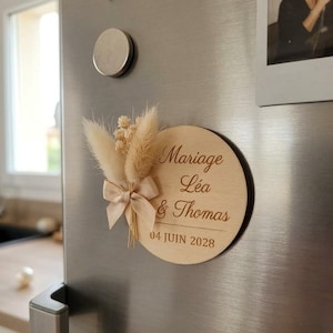 Puede incluir: Un imán redondo de madera con la inscripción "Mariage Léa & Thomas 04 JUIN 2028". Está adornado con un lazo beige y flores secas. El imán está adherido a un refrigerador plateado.
