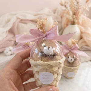 Puede incluir: Una pequeña cesta tejida contiene una esfera transparente con un osito de peluche, adornada con un lazo rosa y flores secas. La cesta tiene una etiqueta que dice "Baptême Louise 14.06.2026".